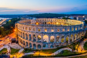 The Pula Arena in Croatia is seen at dusk as a large oval Roman amphitheatre with multiple arcaded tiers and an open inner arena.