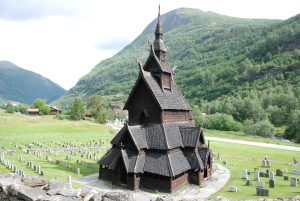 Borgund Stave Church from an elevated perspective with a cemetery and green mountain slopes in the background.