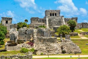 Central stepped structure at Tulum behind walls and smaller stone ruins beneath a blue sky.