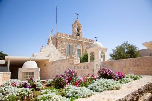Stone building of the Milk Grotto with a bell tower, crosses, small domes and flowers in the foreground.