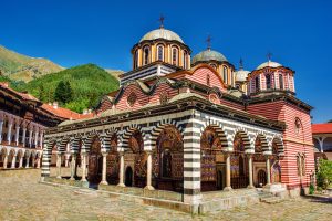 Church at Rila Monastery with black-and-white arcades, colorful frescoes and several domes in front of wooded mountains.
