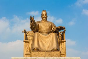 Statue of King Sejong seated frontally on his throne with raised hand and open book against a blue sky